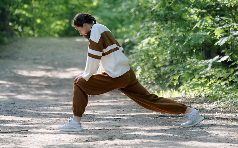 10 - A person doing a side lunge stretch in a forest wearing a cream and brown striped sweater and matching brown sweatpants